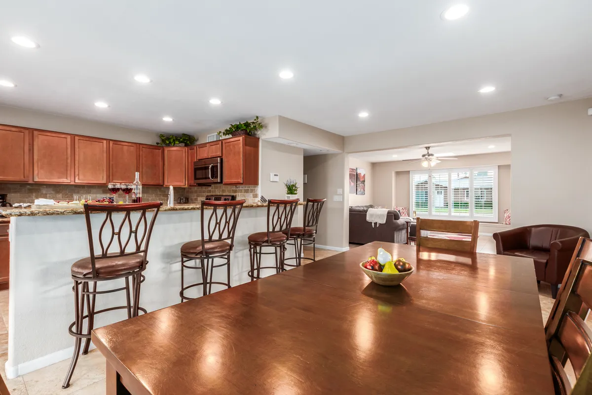 An open-plan kitchen and dining area with a kitchen island, bar stools for casual seating, and a large dining table nearby. Sunlight streams through large windows, uniting the spaces for both casual bites and group meals.