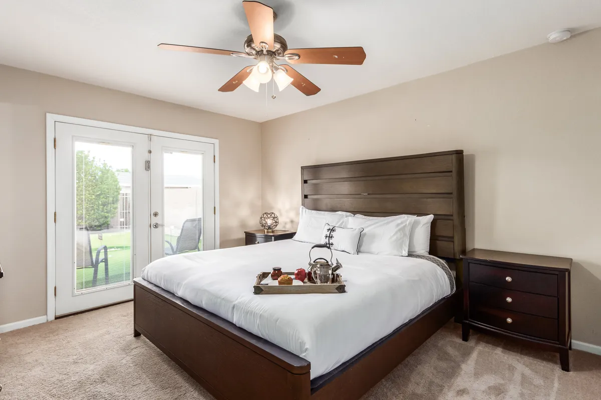 A serene bedroom featuring a bed adorned with a decorative tray, double doors that lead to an outdoor space, a ceiling fan, and soft, neutral decor. Natural light pours in through the doors, creating a peaceful, restful atmosphere.