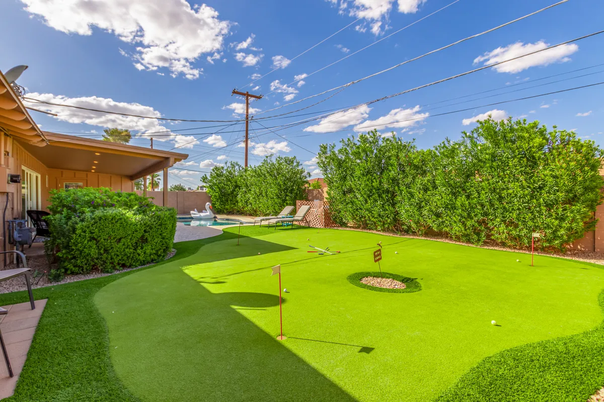 A playful backyard putting green accompanied by green hedges and comfortable lounge chairs under a clear blue sky. The setup is ideal for friendly competitions and outdoor relaxation, blending fun and leisure.