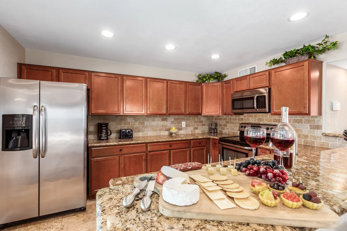 A close-up view of a kitchen island set for entertaining, featuring a granite countertop, a cheese platter, wine glasses, and stainless steel appliances in the background. The setup suggests a lively, welcoming environment for conversation and special moments.