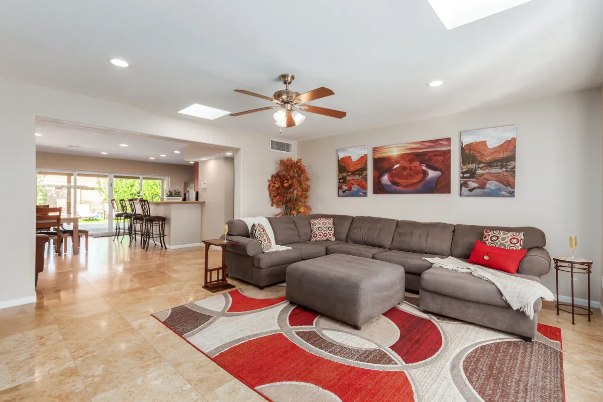 A cozy, open-concept living area with a large sectional sofa, colorful area rug, artistic wall decor, and a glimpse of a dining area in the background. The space is inviting for relaxation, movie nights, or group gatherings.