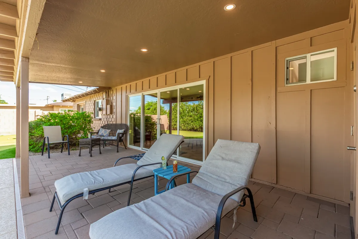 A tranquil backyard setting featuring cushioned lounge chairs grouped around a small blue table, set on a patio just outside sliding glass doors. Lush greenery lines the perimeter, creating a private and inviting atmosphere for gatherings or relaxation.