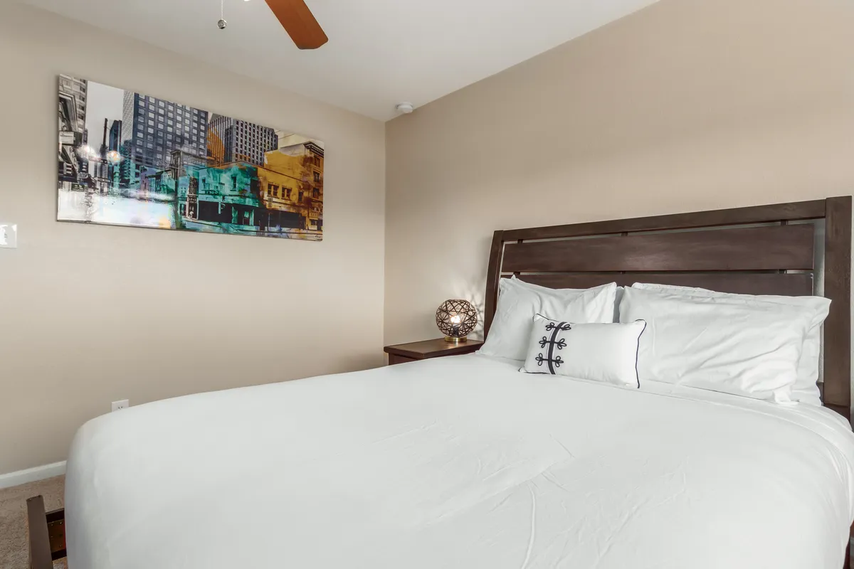 A spacious bedroom with a large bed dressed in crisp white bedding, vibrant wall art, a nightstand with a decorative lamp, and ample natural light coming through a window. The space feels inviting and ideal for relaxation.